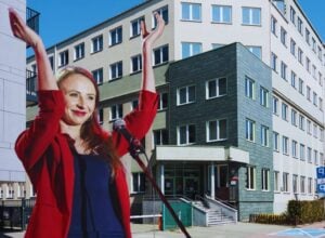 Agnieszka Dziemianowicz-Bąk w czerwonej marynarce, z uniesionymi rękami na tle budynku Państwowej Inspekcji Pracy