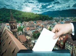 Zdjęcie przedstawia rynek w Badenii-Wirtembergii, mnóstwo kolorowych kamienic, na tym tle doklejona dłoń wrzucająca do urny kartę do głosowania