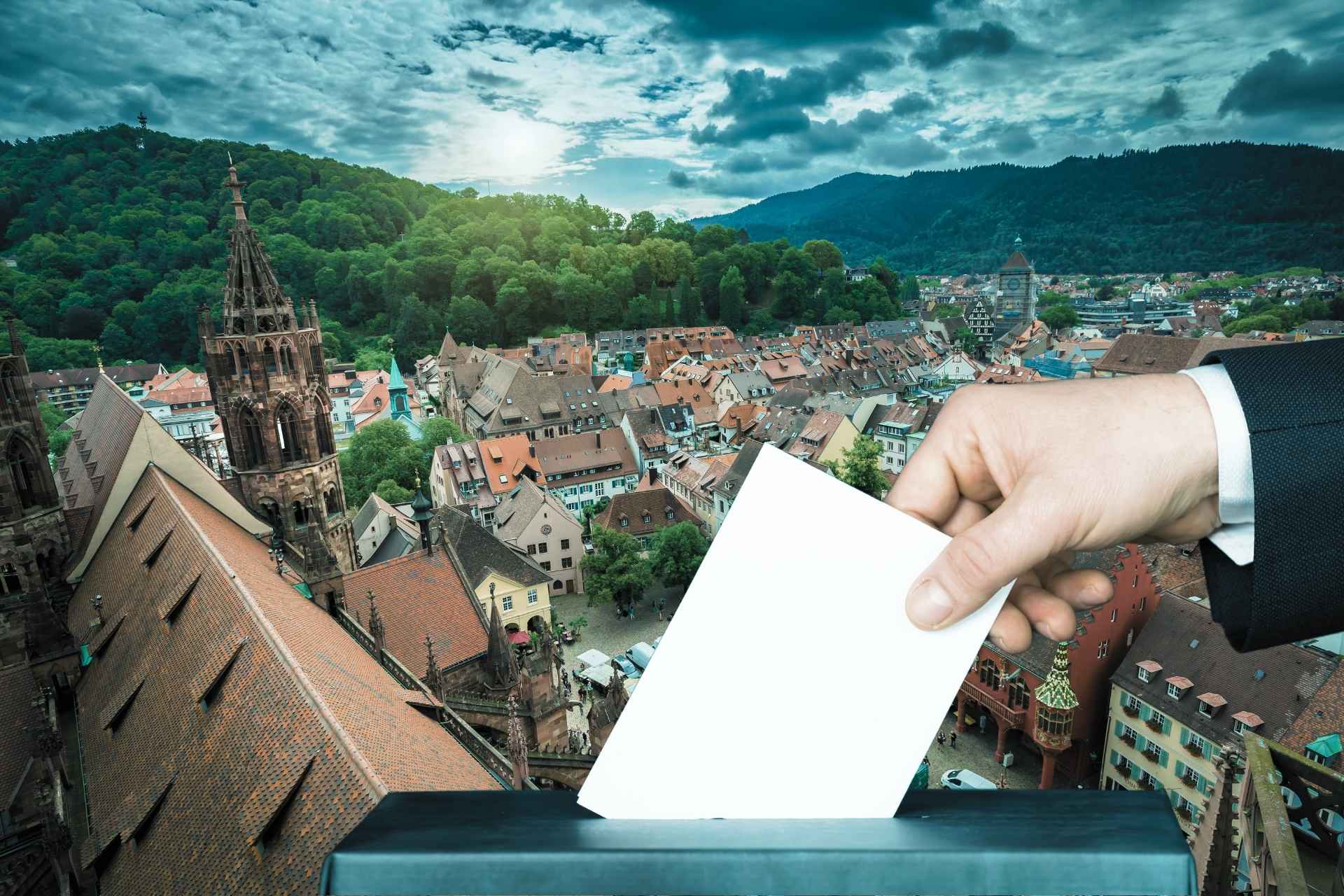 Zdjęcie przedstawia rynek w Badenii-Wirtembergii, mnóstwo kolorowych kamienic, na tym tle doklejona dłoń wrzucająca do urny kartę do głosowania