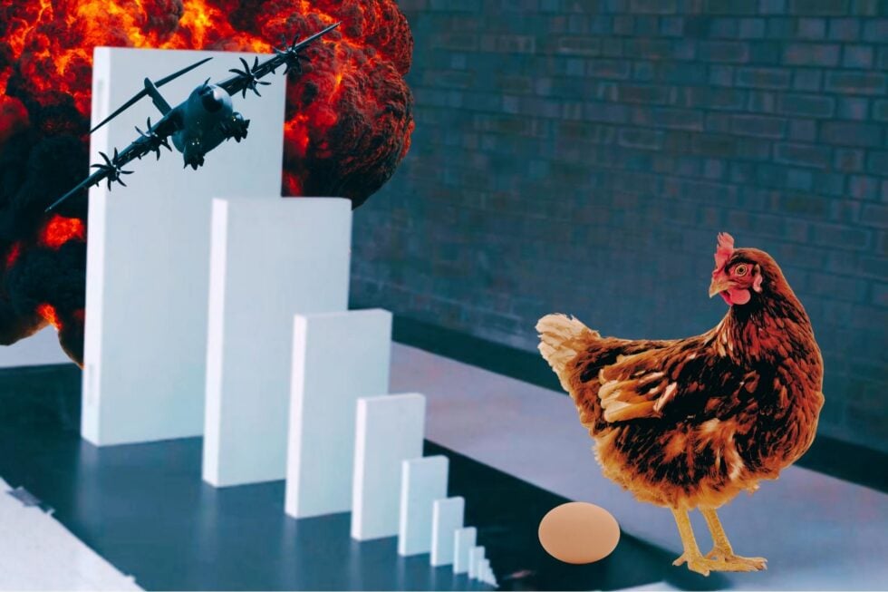 Wielkie białe klocki Domino, przy najmniejszym z przodu stoi kura, pod nią leży jajko. Przy największym klocku widać samolot bojowy, a za nim wybuch.