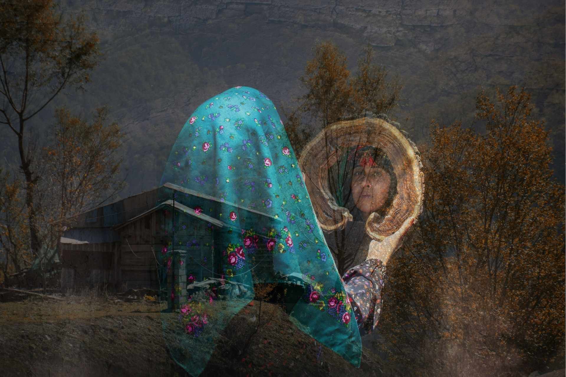 Drewniany domek na pustkowiu, na to nałożony półprzezroczysty obraz młodej irańskiej kobiety w niebieskiej chuście stojącej tyłem, patrzącej w lustro, w którym odbija się jej twarz