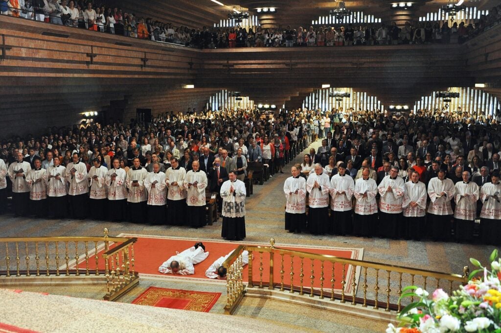 Modły w sanktuarium Torreciudad. Dwóch mężczyzn leży twarza do ziemi w kościele pełnym księzy. 