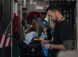 Ratownik medyczny wkłada rękawice, w tle pacjent przypięty do noszy i szarpiący się z sanitariuszem. Kadr z serialu The Pitt