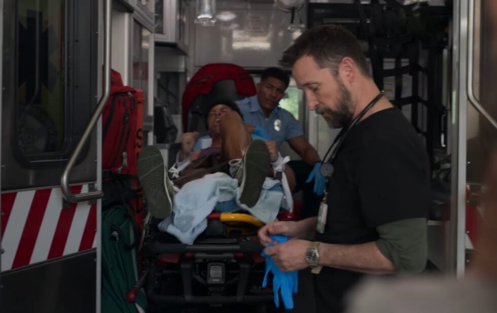 Ratownik medyczny wkłada rękawice, w tle pacjent przypięty do noszy i szarpiący się z sanitariuszem. Kadr z serialu The Pitt