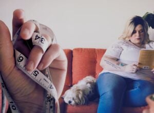 Blondynka z nadwagą czytająca książkę siedząc na kanapie, obok niej leży mały kudłaty pies, po lewej stronie doklejona jest dłoń trzymająca centymetr