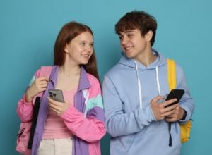 Na niebieskim tle nastoletnia dziewczyna w różowej kurtce i chłopak w niebieskiej bluzie trzymają w rękach telefony, uśmiechając się i spoglądając na siebie