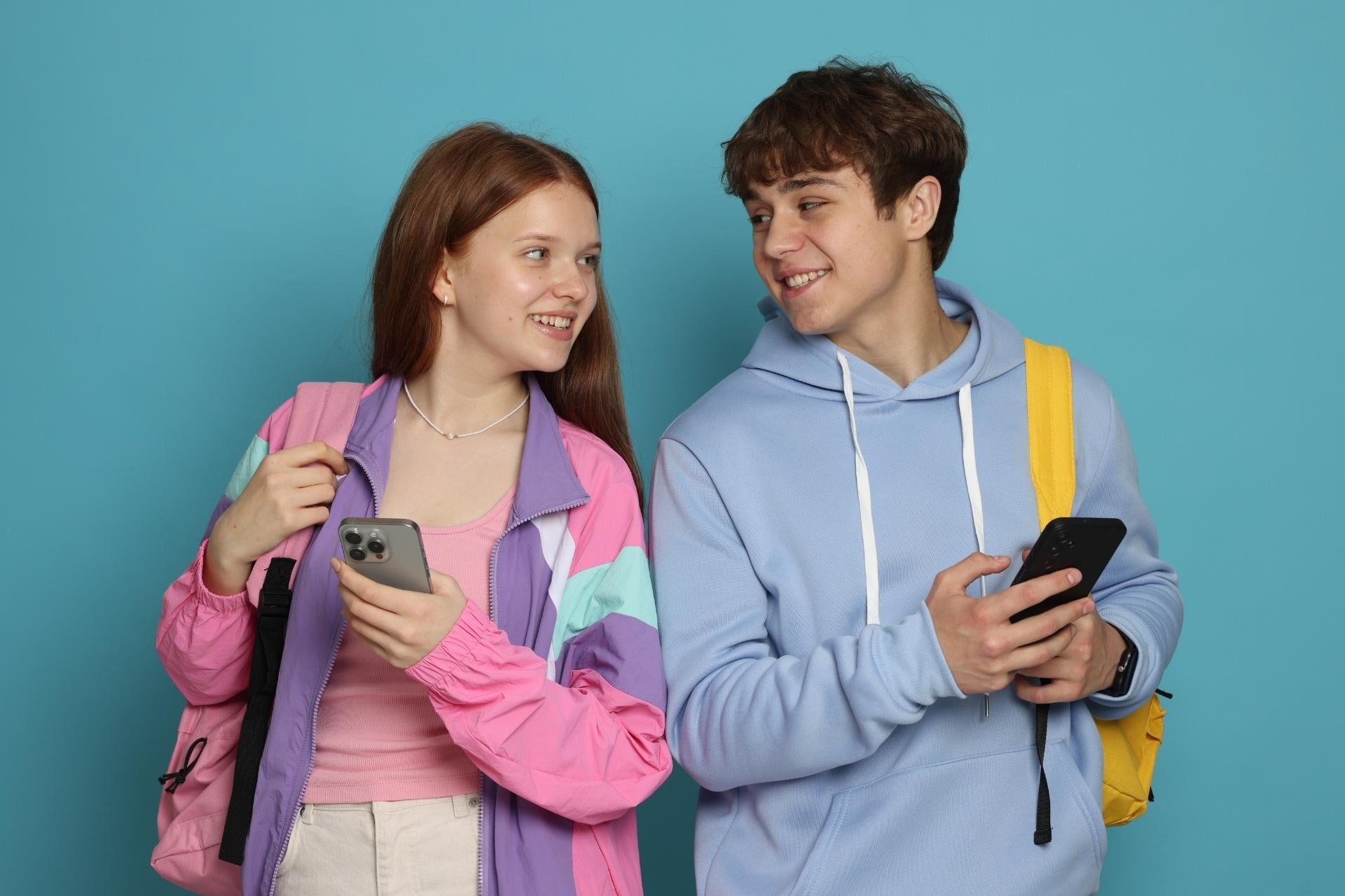 Na niebieskim tle nastoletnia dziewczyna w różowej kurtce i chłopak w niebieskiej bluzie trzymają w rękach telefony, uśmiechając się i spoglądając na siebie