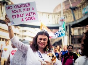 Uśmiechnięta Harish Iyer w białym, tradycyjnym indyjskim stroju i z pluszowym pieskiem w ręce, w prawej ręce unosi białą kartkę z hasłem „No gender role, just a stray with a soul”