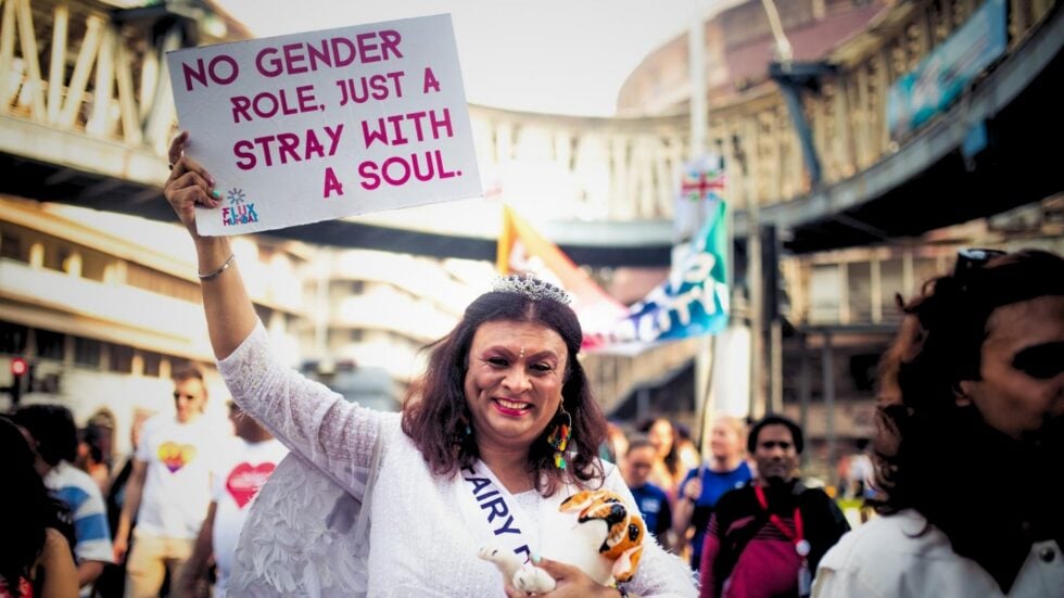 Uśmiechnięta Harish Iyer w białym, tradycyjnym indyjskim stroju i z pluszowym pieskiem w ręce, w prawej ręce unosi białą kartkę z hasłem „No gender role, just a stray with a soul”