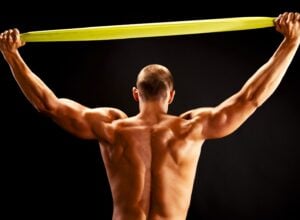Bardzo umięśniony mężczyzna bez koszulki widoczny od tyłu, rozciągający nad głową żółtą taśmę