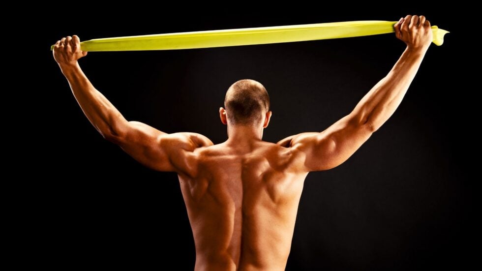 Bardzo umięśniony mężczyzna bez koszulki widoczny od tyłu, rozciągający nad głową żółtą taśmę
