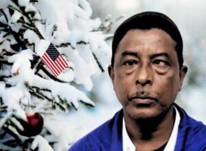 Po prawej Nurul Amin Shah Alam, mężczyzna w średnim wieku z widoczną wadą wzroku, w tle śnieg, po lewej stronie kawałek choinki z wetkniętą flagą USA