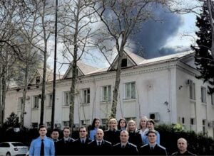 Grupa prokuratorów i prokuratorek stojąca na tle dużego białego budynki, za którym z nieba unosi się czarny dym