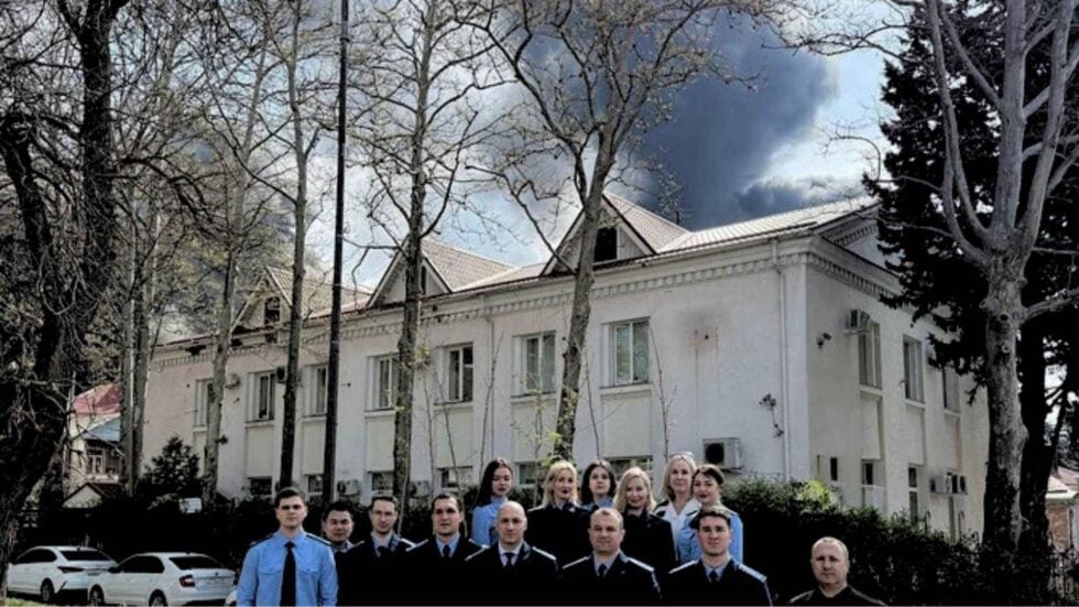 Grupa prokuratorów i prokuratorek stojąca na tle dużego białego budynki, za którym z nieba unosi się czarny dym