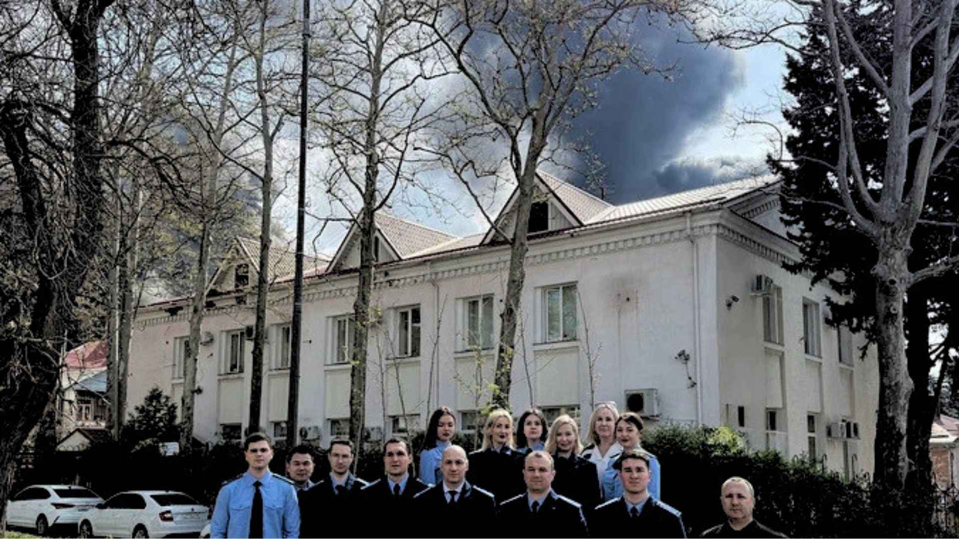 Grupa prokuratorów i prokuratorek stojąca na tle dużego białego budynki, za którym z nieba unosi się czarny dym