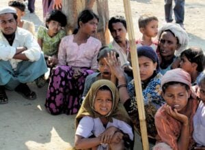 Kilkanaście osób z grupy Rohingya, dzieci i dorośli, siedzący na gołej ziemi, wszyscy mają smutny wyraz twarzy, część patrzy w obiektyw