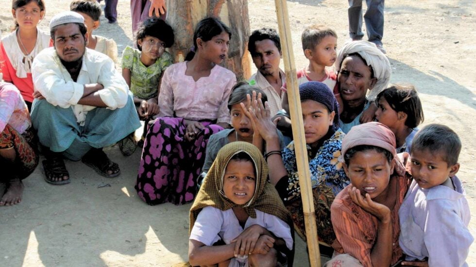 Kilkanaście osób z grupy Rohingya, dzieci i dorośli, siedzący na gołej ziemi, wszyscy mają smutny wyraz twarzy, część patrzy w obiektyw