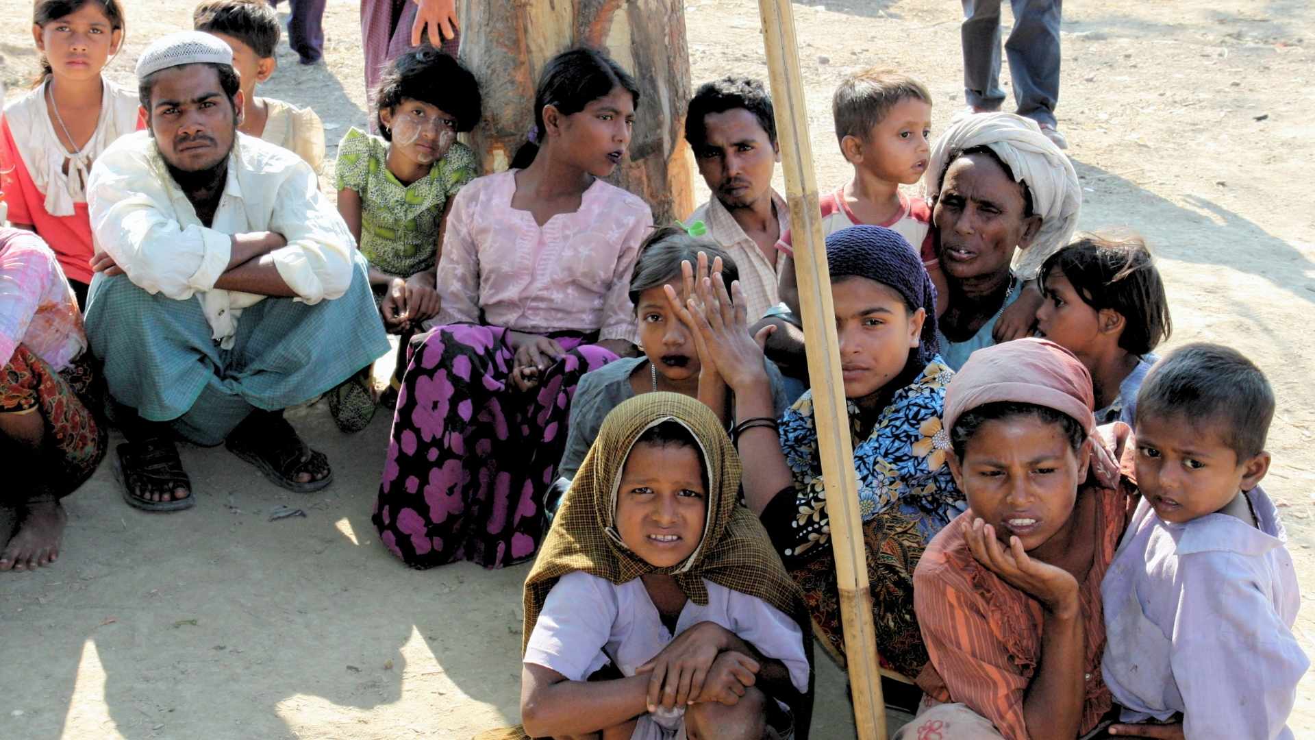 Kilkanaście osób z grupy Rohingya, dzieci i dorośli, siedzący na gołej ziemi, wszyscy mają smutny wyraz twarzy, część patrzy w obiektyw