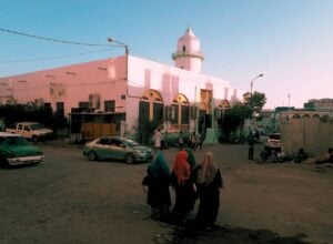 Widok na meczet w Dżibuti z niewielkim minaretem, pod nim stoją stare samochody, w centrum zdjęcia znajdują się trzy odwrócone tyłem kobiety w hidżabach, idące w kierunku meczetu