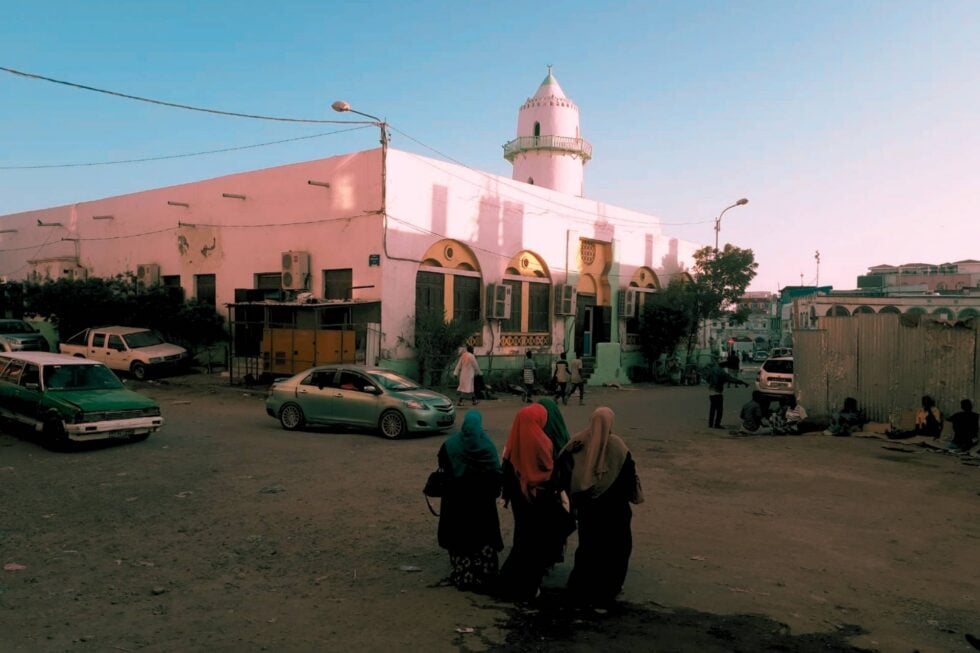 Widok na meczet w Dżibuti z niewielkim minaretem, pod nim stoją stare samochody, w centrum zdjęcia znajdują się trzy odwrócone tyłem kobiety w hidżabach, idące w kierunku meczetu