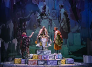 Stage performance with three costumed actors on a stack of oversized money blocks; central figure sits on a throne-like chair while others converse.