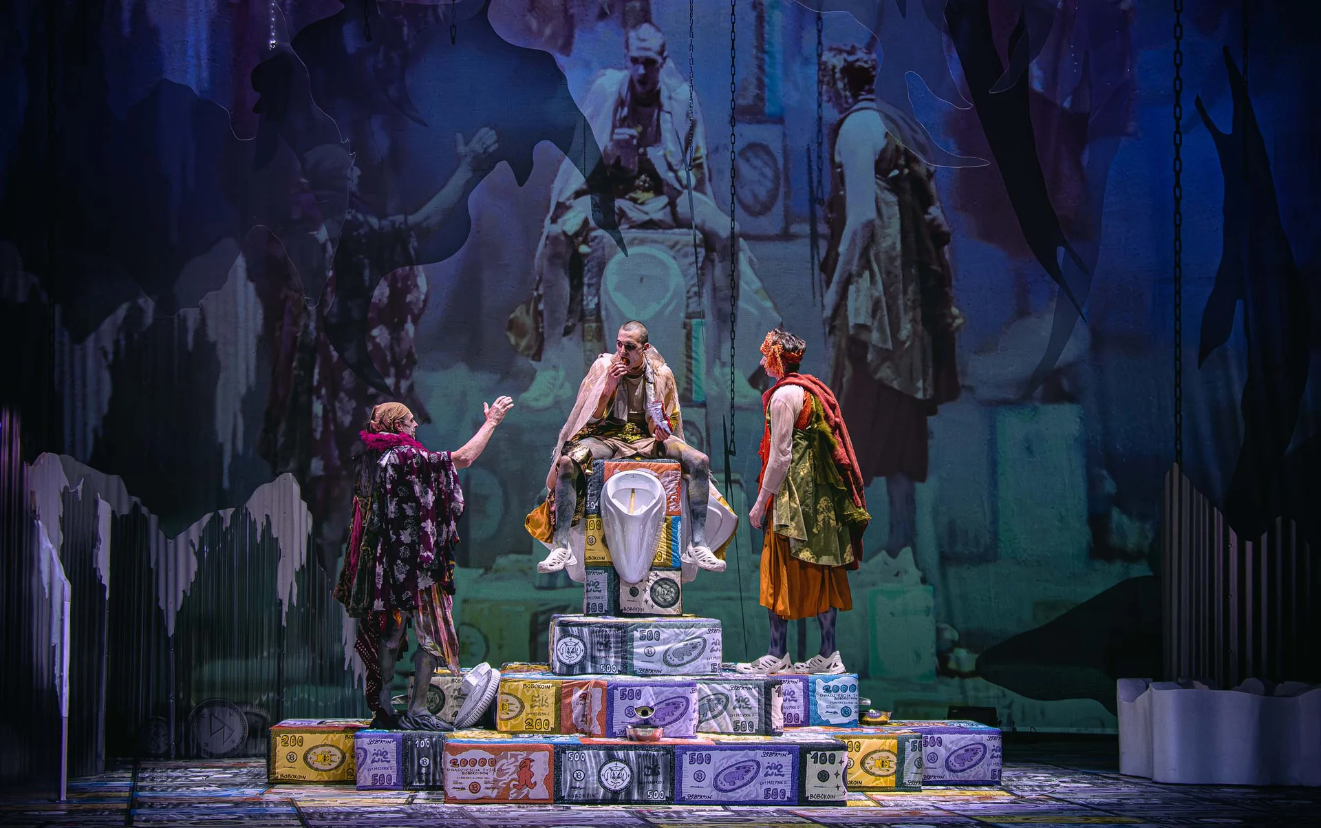 Stage performance with three costumed actors on a stack of oversized money blocks; central figure sits on a throne-like chair while others converse.