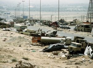 Desert highway strewn with wrecked cars, buses, and debris after a disaster; city in the hazy distance on the horizon.