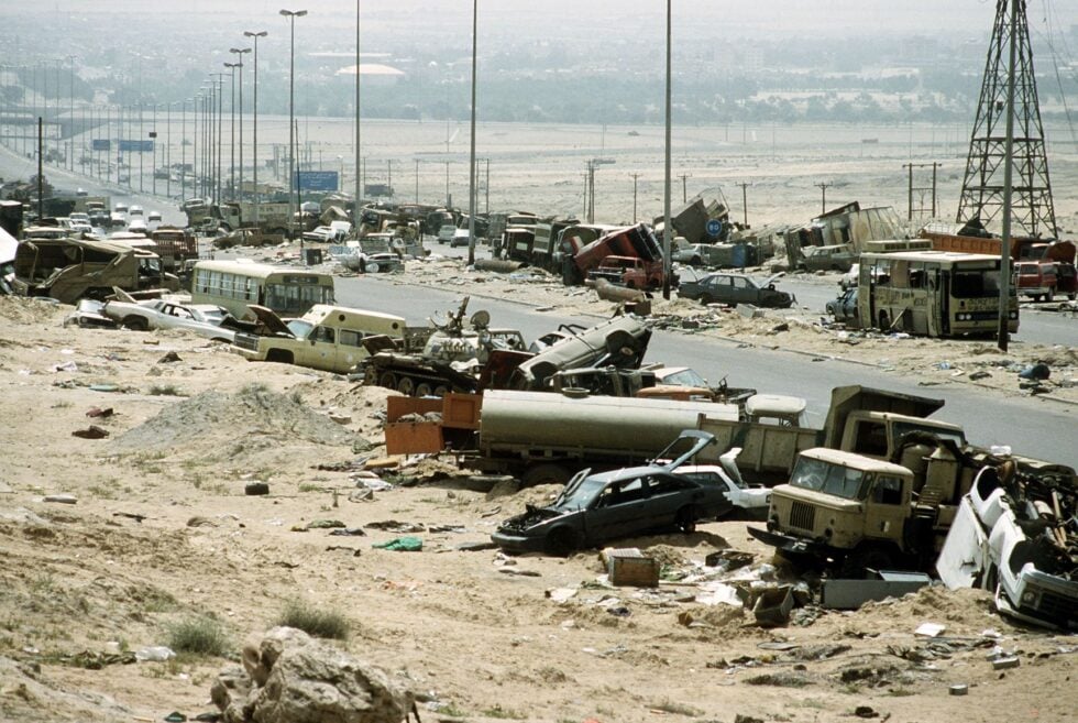 Desert highway strewn with wrecked cars, buses, and debris after a disaster; city in the hazy distance on the horizon.