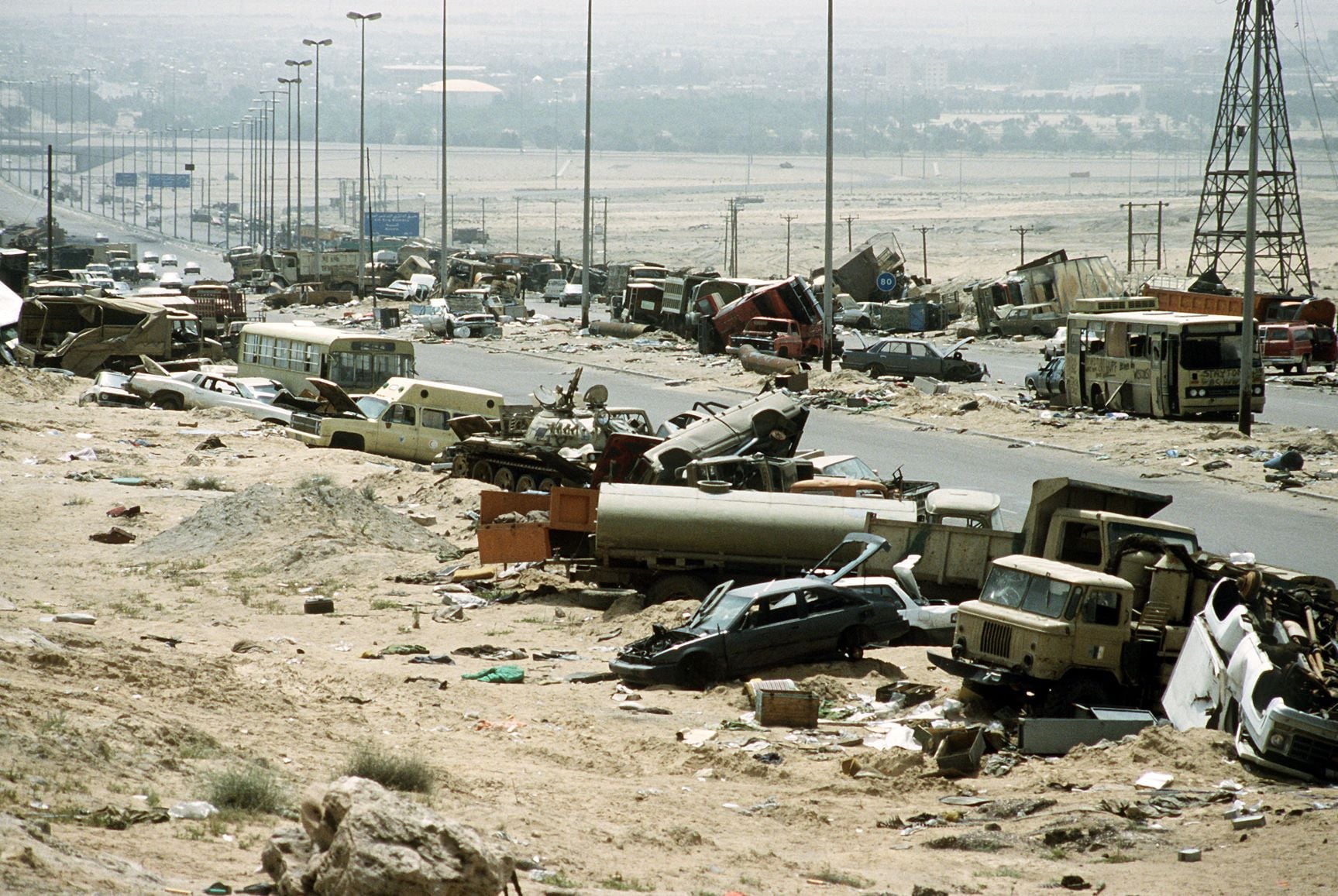 Desert highway strewn with wrecked cars, buses, and debris after a disaster; city in the hazy distance on the horizon.