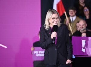 Woman in a black suit smiles as she speaks into a handheld microphone on a purple-lit stage.