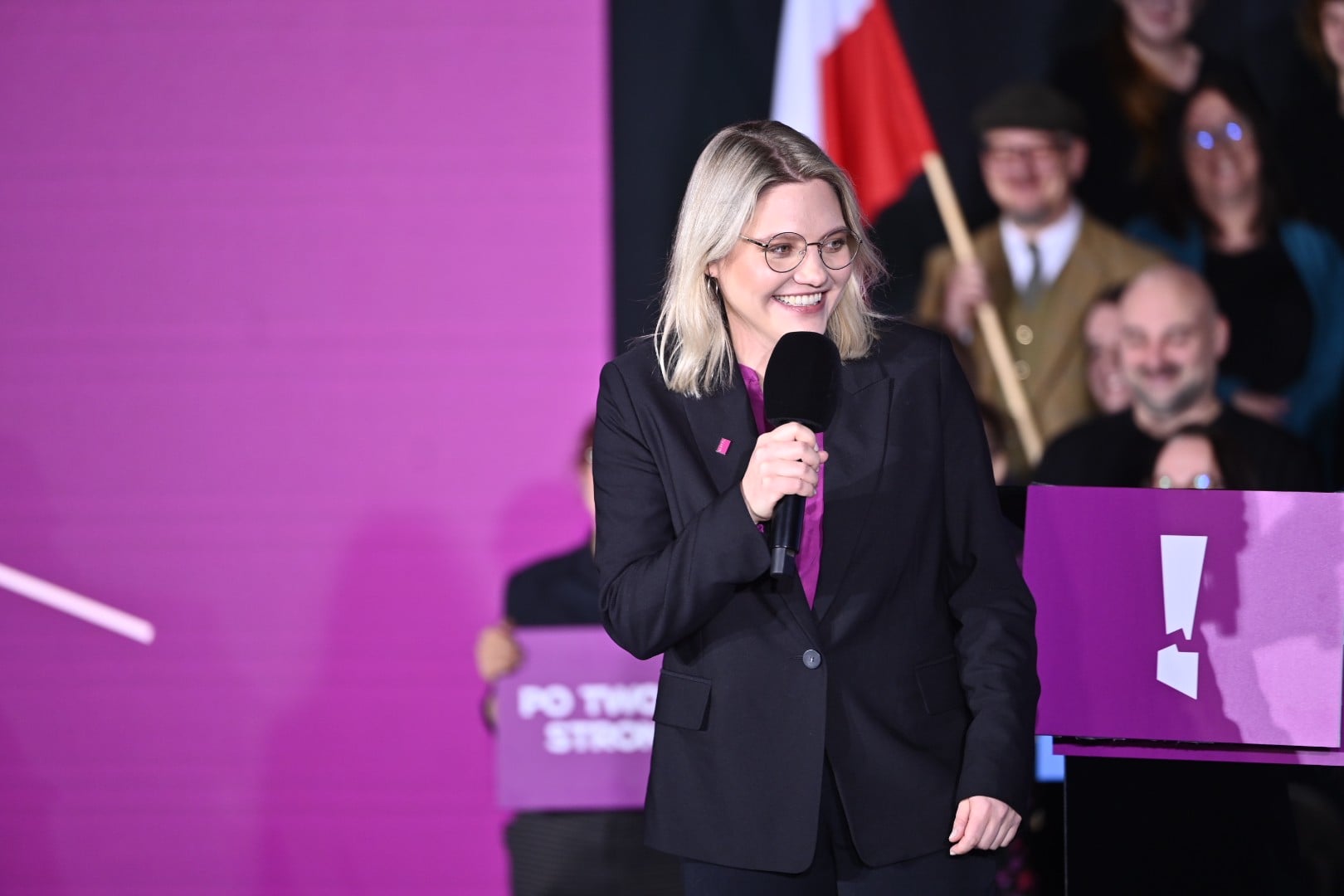 Woman in a black suit smiles as she speaks into a handheld microphone on a purple-lit stage.