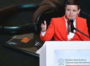 A woman in a red blazer speaks into a microphone at a podium during a formal event for the Polish Ministry of Culture and National Heritage, with large headphones in the background on a table nearby.