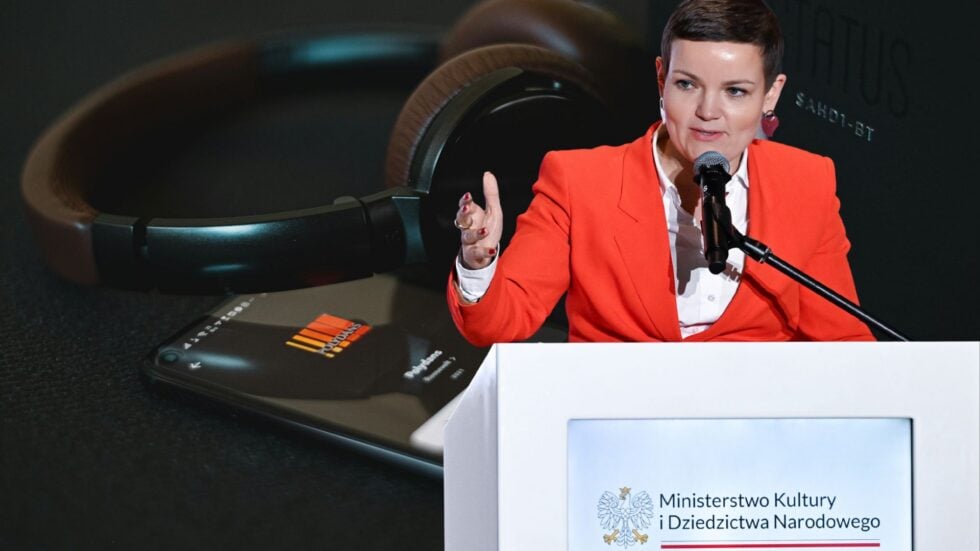 A woman in a red blazer speaks into a microphone at a podium during a formal event for the Polish Ministry of Culture and National Heritage, with large headphones in the background on a table nearby.
