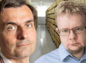 Two men pose in a storage warehouse; tall shelves with yellow boxes line the aisle behind them.