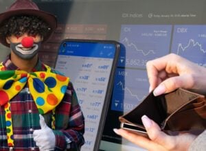 Clown with red nose and painted smile wearing a colorful bow tie and plaid shirt, posing beside stock market screens.