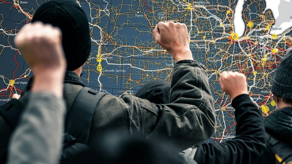 Group of people examining a large road map with many colored routes and city labels on a wall screen/board.