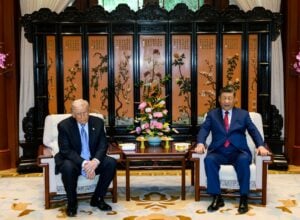 Two men in suits seated on armchairs in an ornate room with a lacquered screen and pink orchid arrangements behind them.