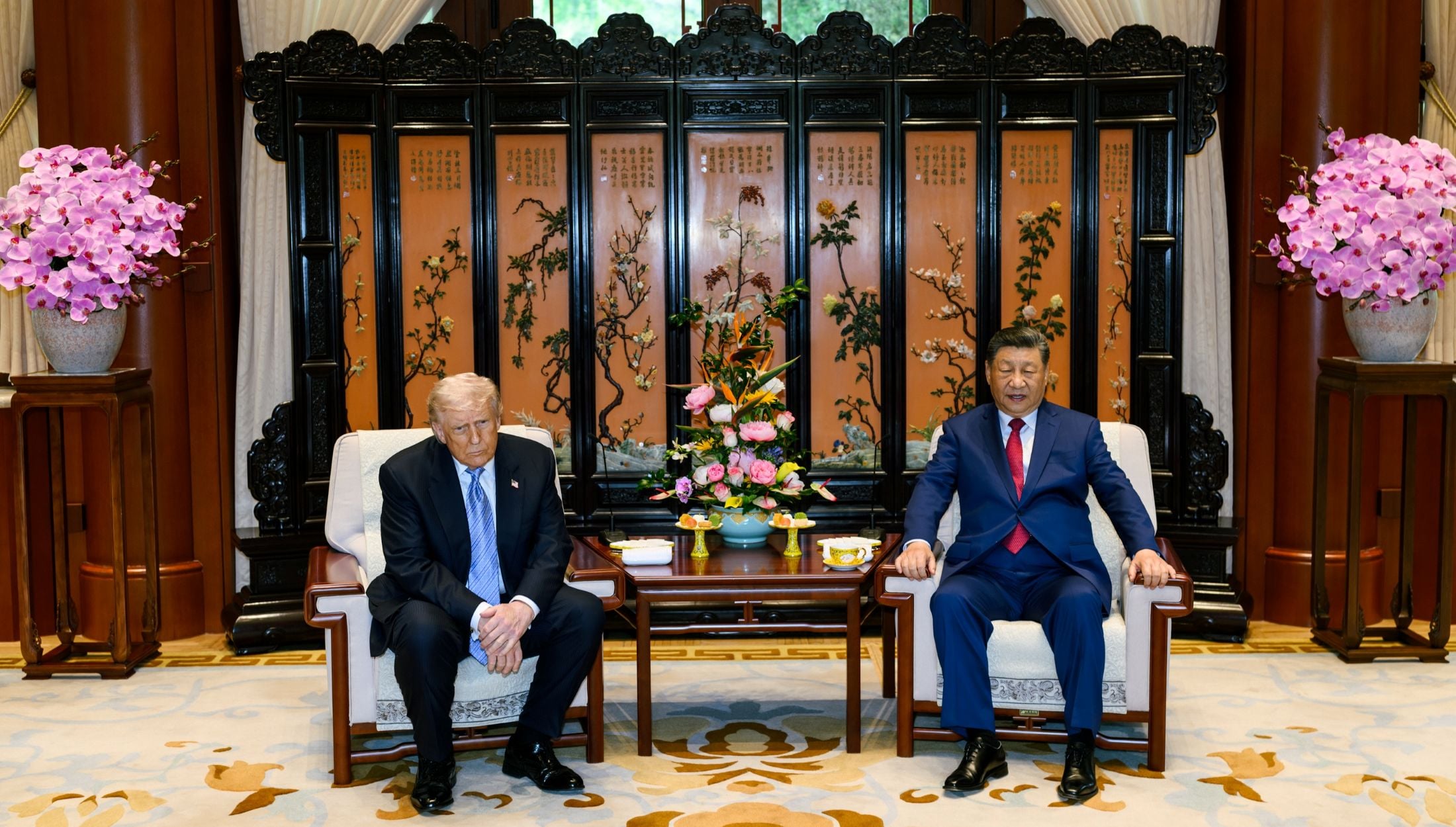 Two men in suits seated on armchairs in an ornate room with a lacquered screen and pink orchid arrangements behind them.