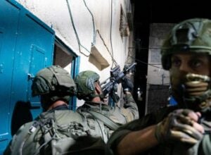 Soldiers in camouflage gear and helmets aiming rifles in a narrow alley at night beside blue doors