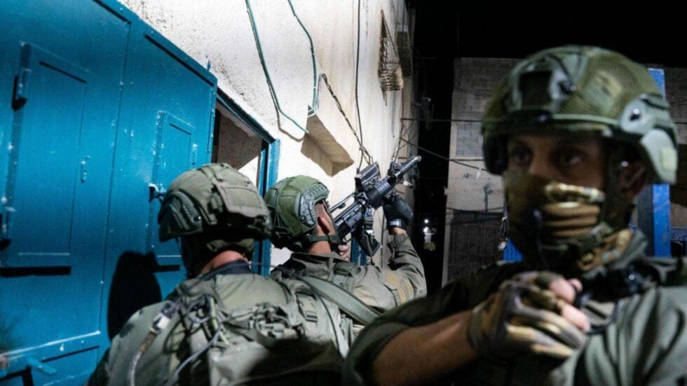 Soldiers in camouflage gear and helmets aiming rifles in a narrow alley at night beside blue doors