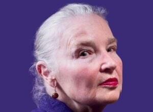 Close-up portrait of an older woman with white hair and red lipstick against a purple background.