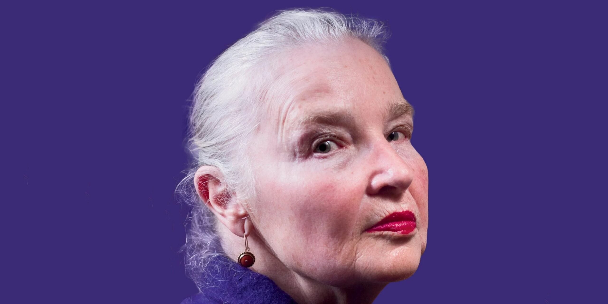 Close-up portrait of an older woman with white hair and red lipstick against a purple background.
