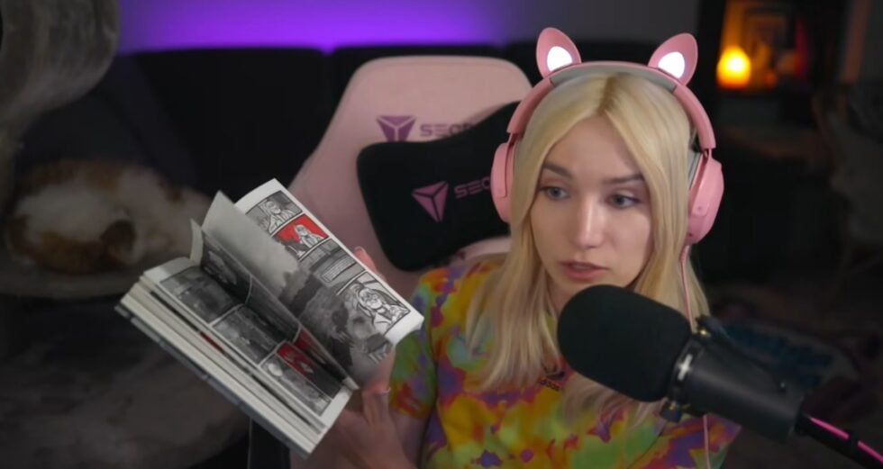 Female streamer with pink cat-ear headset reading a black-and-white comic book into a microphone in a dimly lit room with purple backlighting and a pink gaming chair behind her.