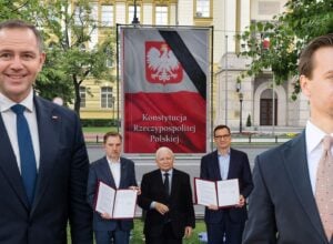 Five men in suits outdoors posing beside a banner that reads Konstytucja Rzeczypospolitej Polskiej near a government-style building.