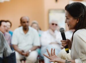 Side-profile of a woman speaking into a microphone at an event, with a blurred audience in the background