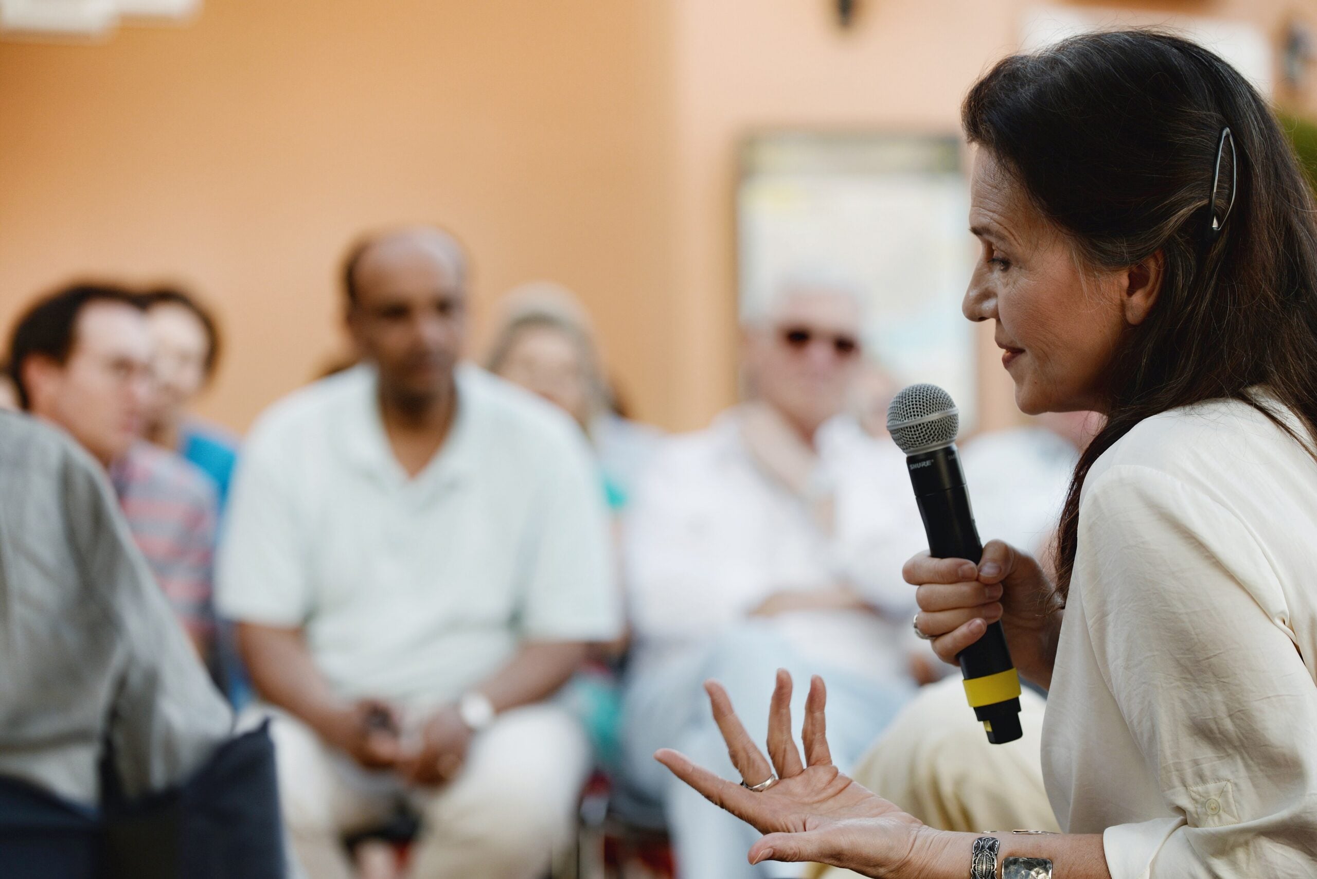 Side-profile of a woman speaking into a microphone at an event, with a blurred audience in the background