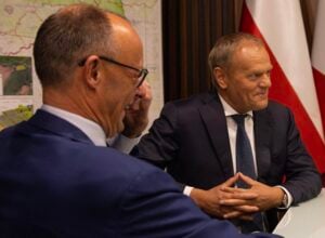 Two businessmen in suits sit at a table during a meeting; the man on the right smiles, hands folded on the table, with a map and flag in the background.
