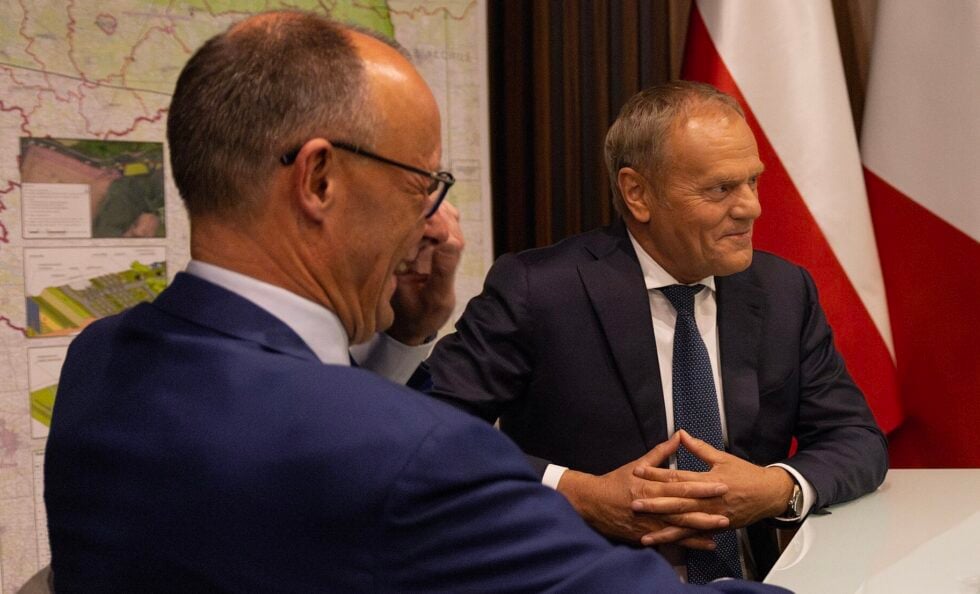 Two businessmen in suits sit at a table during a meeting; the man on the right smiles, hands folded on the table, with a map and flag in the background.