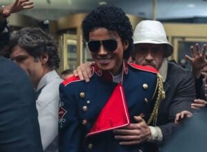 Kadr z filmu „Michael” (2026) przedstawiający Michaela Jacksona wśród ludzi, w eleganckim granatowo-czerwonym mundurze ze złotymi guzikami, obejmowanego od tyłu przez mężczyznę w kapeluszu
