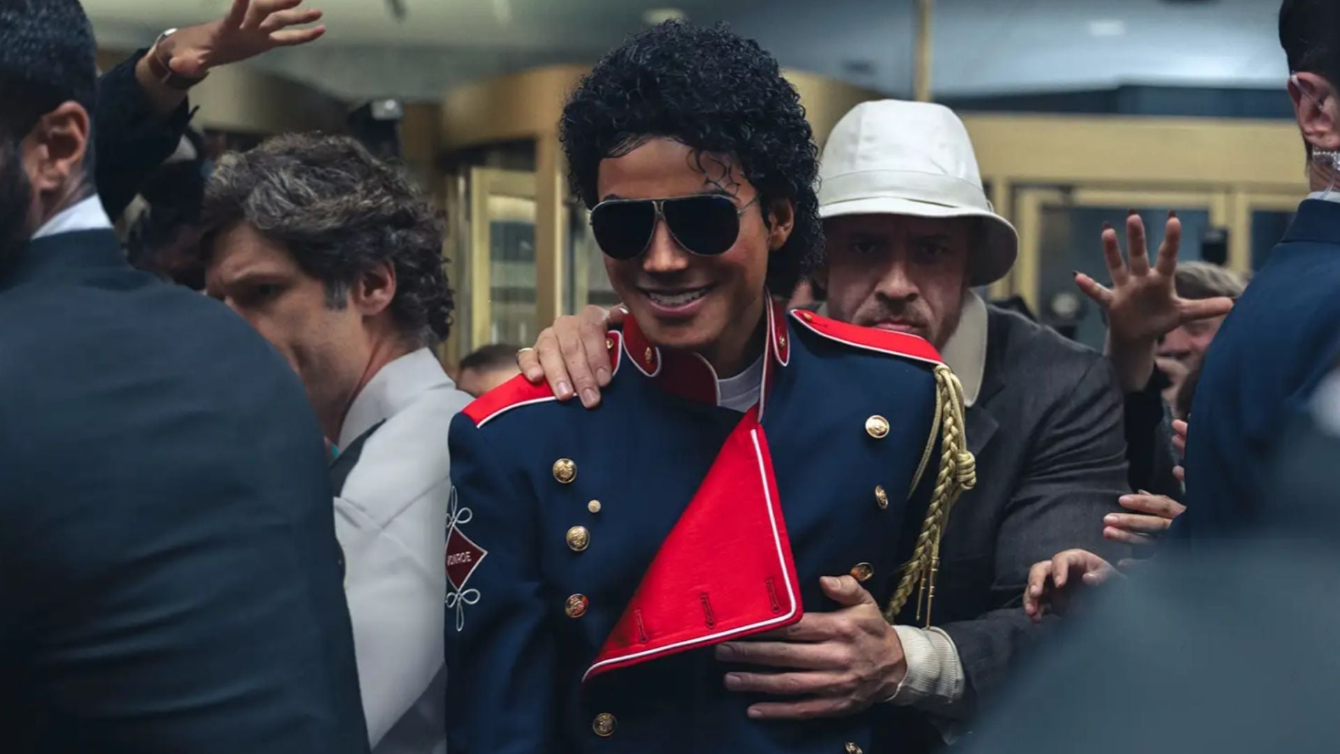 Kadr z filmu „Michael” (2026) przedstawiający Michaela Jacksona wśród ludzi, w eleganckim granatowo-czerwonym mundurze ze złotymi guzikami, obejmowanego od tyłu przez mężczyznę w kapeluszu
